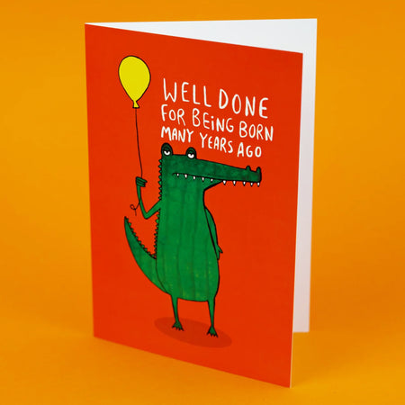 a red A6 greeting card with a illustration of an unimpressed looking green crocodile holding a yellow balloon with white text above it that reads 'well done for being born many years ago'. Designed by Katie Abey in the UK. Available on Rebel Romance