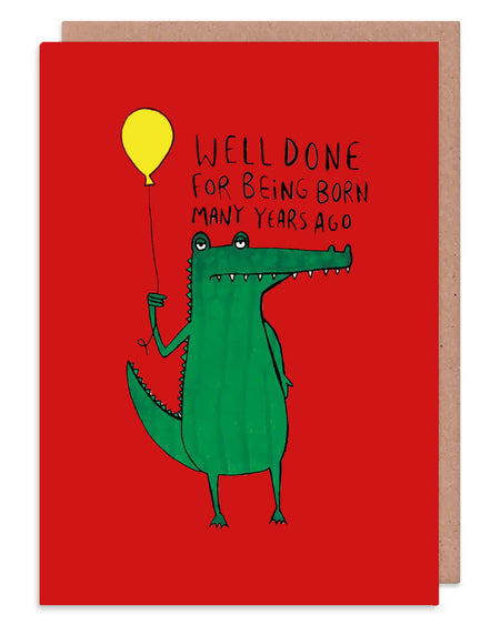 a red A6 greeting card with a illustration of an unimpressed looking green crocodile holding a yellow balloon with white text above it that reads 'well done for being born many years ago'. Designed by Katie Abey in the UK. Available on Rebel Romance
