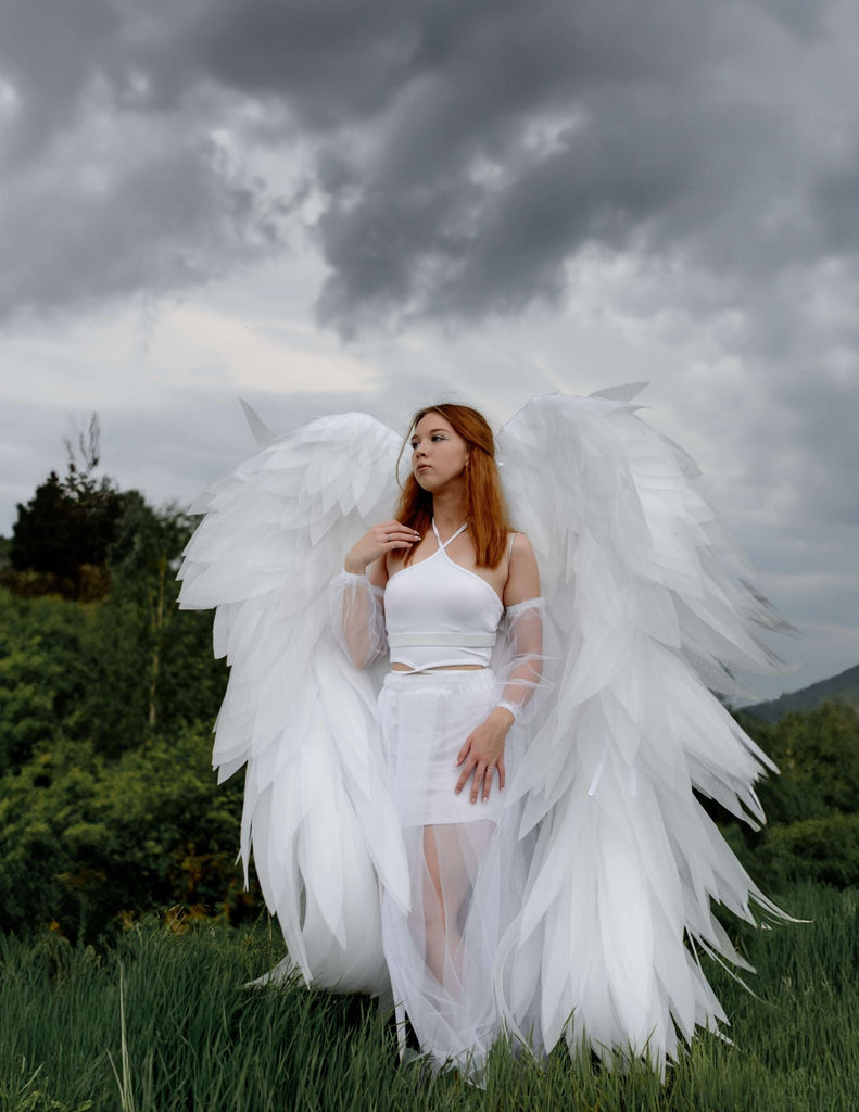 Ksenia dressed as an angel outdoors against a grey sky. She is all in white with giant wings