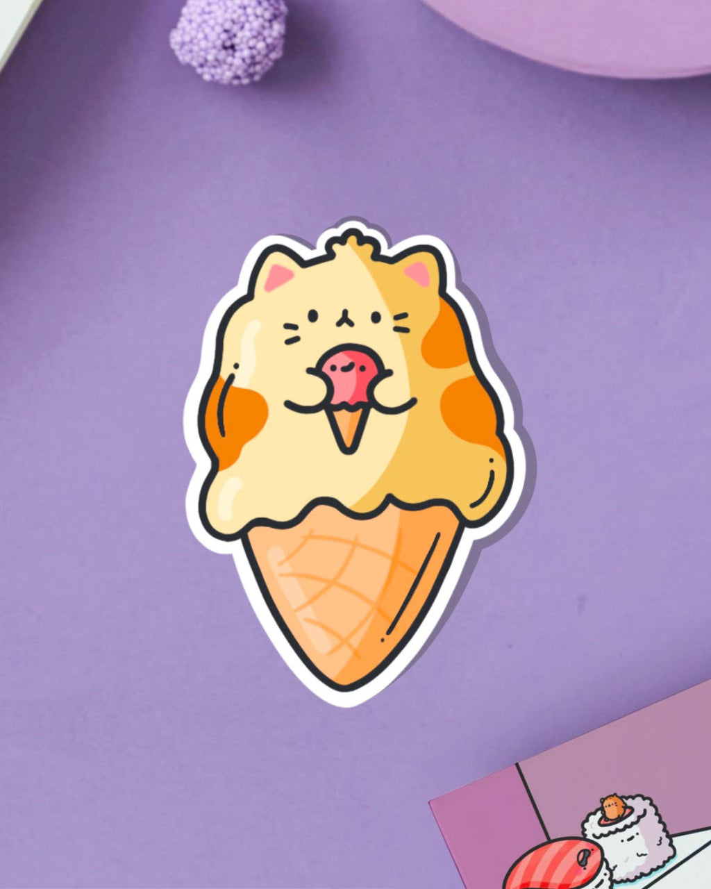 Cute Toastedink vinyl sticker featuring an orange and cream cartoon cat shaped like an ice cream cone, placed on a lilac surface with a purple candy and pastel decor.