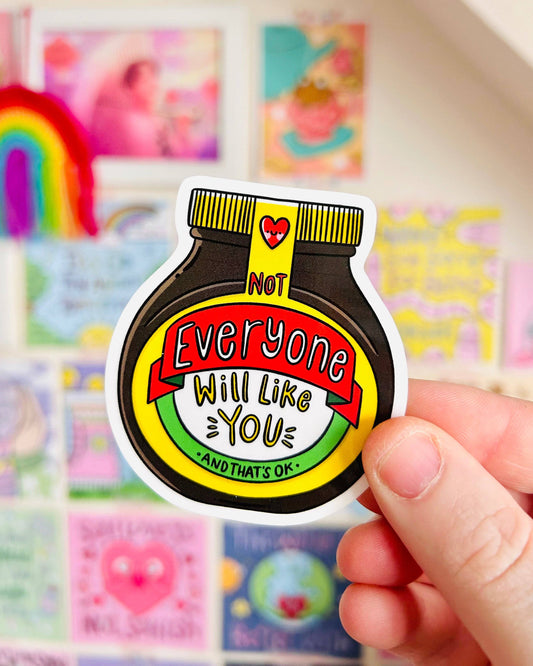 A Marmite-inspired vinyl sticker reading “Not Everyone Will Like You – And That’s OK” is held up in front of a colourful wall of uplifting art. Designed by Kellylou and sold by Rebel Romance, a UK business celebrating alternative style and self-acceptance.