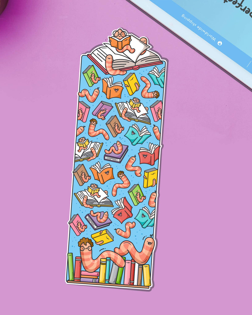 Colourful bookworm bookmark by Toastedink, sold by Rebel Romance, featuring cheerful cartoon worms with glasses surrounded by smiling books on a blue background. Displayed on a purple backdrop.