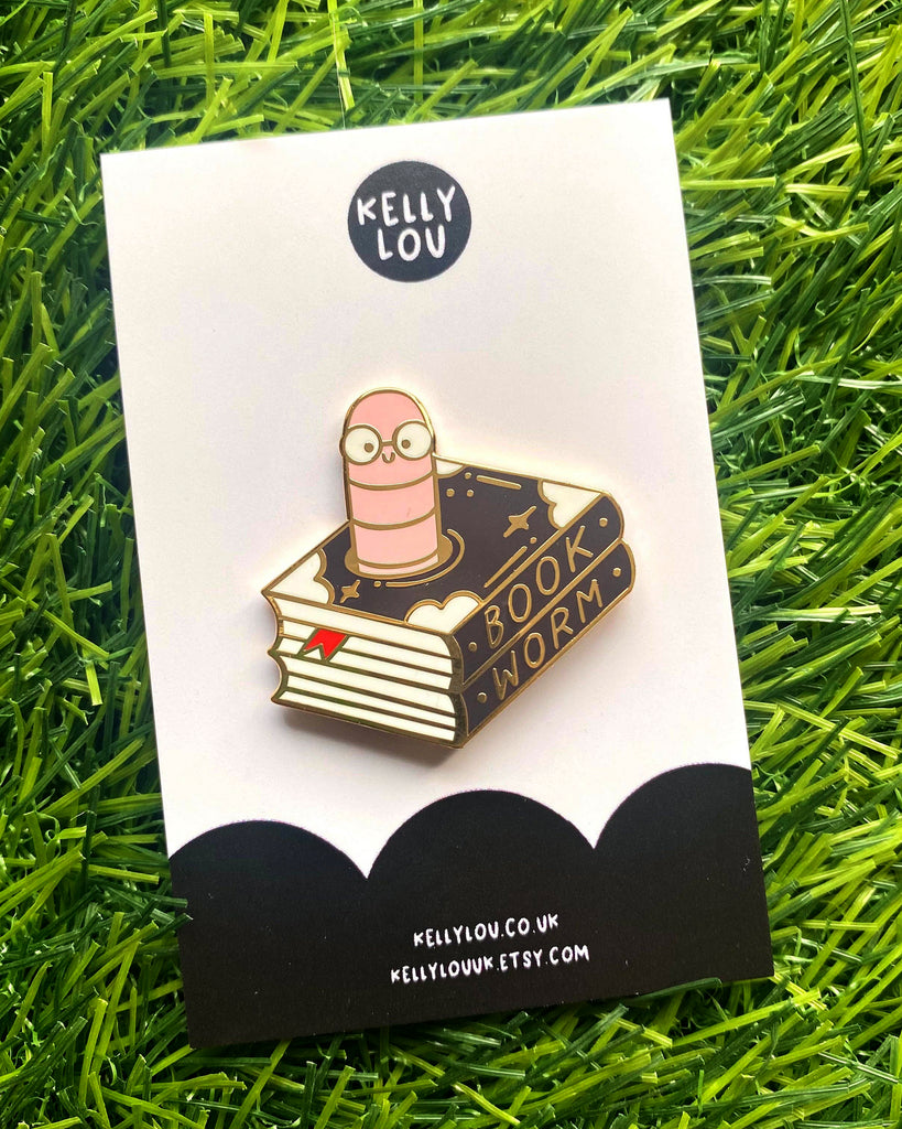 Flat-lay of the Kelly Lou Bookworm Enamel Pin on a Rebel Romance backing card, shown on artificial grass. The gold hard enamel pin features a cartoon pink worm with glasses on a starry black book labelled "Book Worm". Sold by Rebel Romance, a UK small business offering unique alternative fashion, homeware, and accessories for millennials.