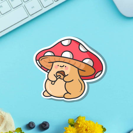 A cute cartoon-style vinyl sticker of a chubby tan mushroom character with a red and white spotted cap, holding a smaller mushroom in its hands. Displayed on a blue background beside a teacup and flower. Waterproof, glossy finish – ideal for decorating everyday items.