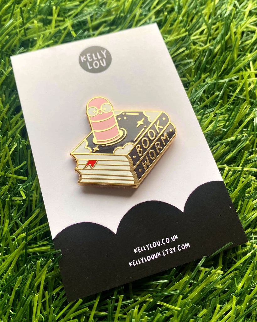 Close-up of the Bookworm Enamel Pin by Kelly Lou, featuring a cute pink worm with glasses popping out of a black and gold book with "Book Worm" written on the spine. Displayed on a branded backing card from Rebel Romance, a UK small business celebrating alternative fashion and quirky gifts. The pin lies on a patch of artificial grass, capturing a fun and playful vibe.