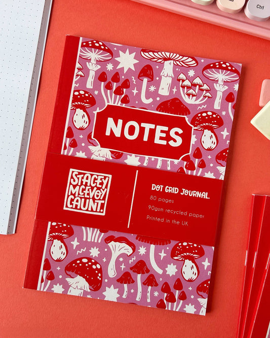 Overhead view of a Stacey McEvoy Caunt mushroom notebook by Rebel Romance. The colourful pink and red cover is decorated with magical mushroom illustrations, and the band states it's printed in the UK on recycled paper.