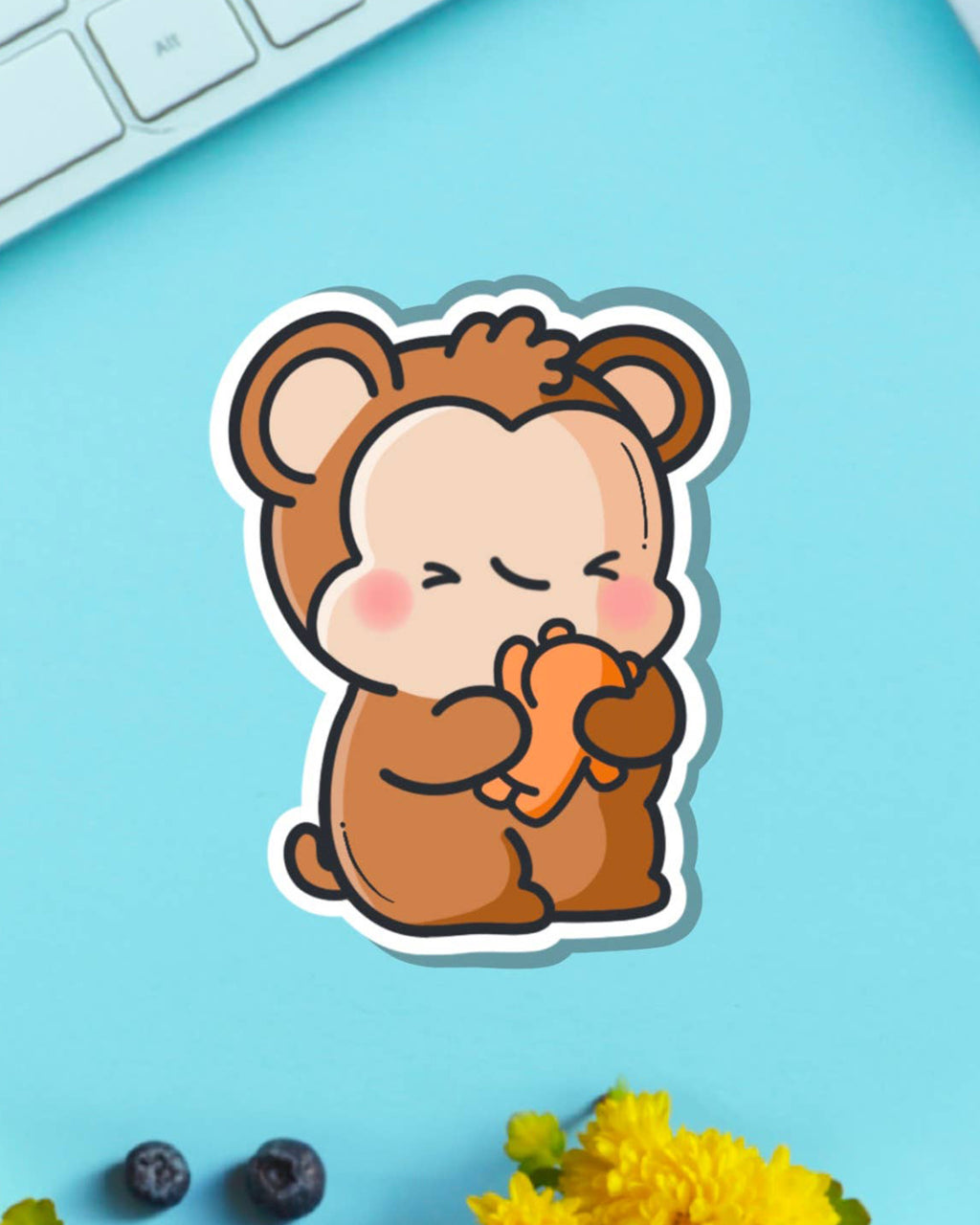 Cute vinyl sticker featuring a cartoon monkey hugging an orange teddy bear, designed by Toastedink. Photographed on a pastel blue background with flowers and keyboard details. Sold by Rebel Romance, a UK brand celebrating quirky and alternative style.