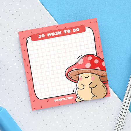 A square sticky note pad with a red border and grid paper design, featuring a cute, blushing mushroom character with a red and white spotted cap in the bottom right corner. The top header reads “So Mush To Do” in bold white text. A fun and functional memo pad for everyday reminders.