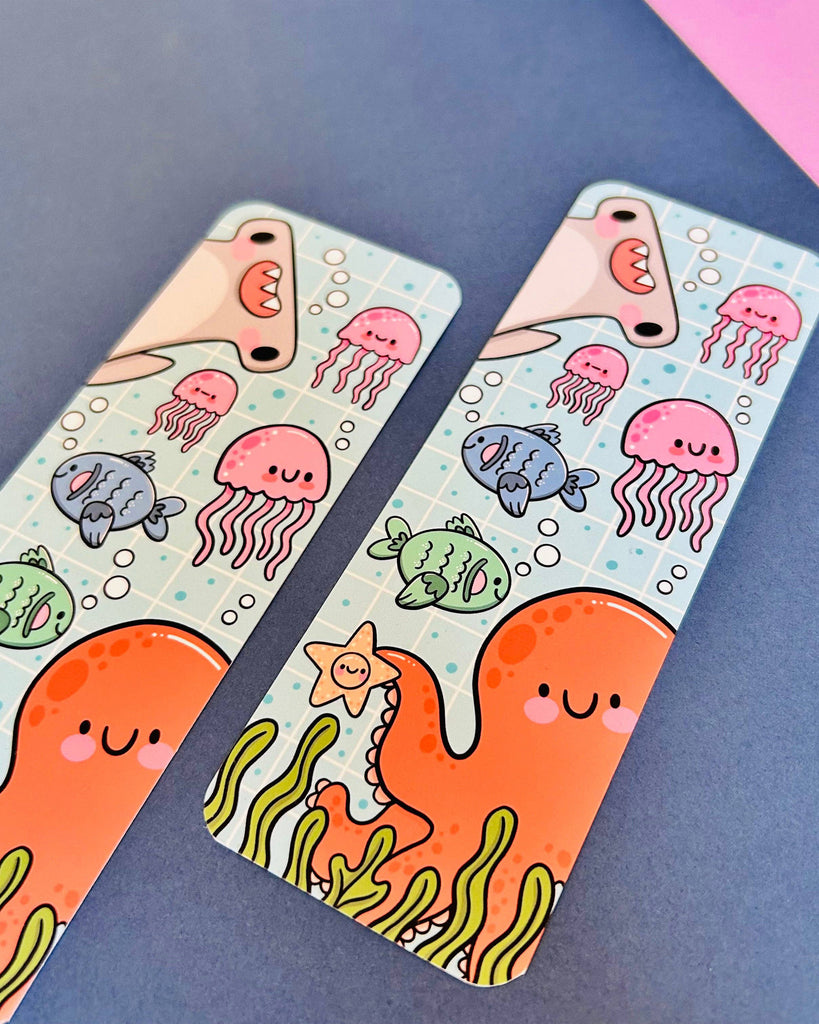 Two Under The Sea bookmarks by Kelly Lou for Rebel Romance shown side-by-side on a blue background with pink accents. Features adorable sea creatures including a smiling octopus, jellyfish, cartoon fish, seaweed, and a goofy shark on a soft blue grid backdrop. Printed on soft-touch velvet laminated card. A cheerful and kawaii accessory for readers, students, and lovers of cute stationery.