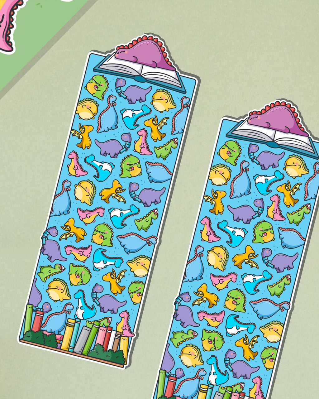 Toastedink's dinosaurus bookmark available at Rebel Romance, with a joyful pattern of rainbow-coloured dinosaurs and a pink dino perched on an open book. Shown against a pale green backdrop.