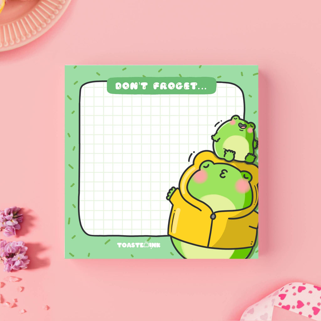 A square frog-themed sticky note pad with a green grid background, featuring a chubby frog in a yellow raincoat being kissed by a smaller frog. The top reads “Don’t Froget…” in a white font on a green tab. Perfect for cute reminders and dopamine-fuelled planning.