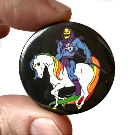 A high-gloss 38mm pin badge featuring Skeletor riding a rainbow-maned unicorn with a black background. A quirky pop culture-inspired design by Bite Your Granny.