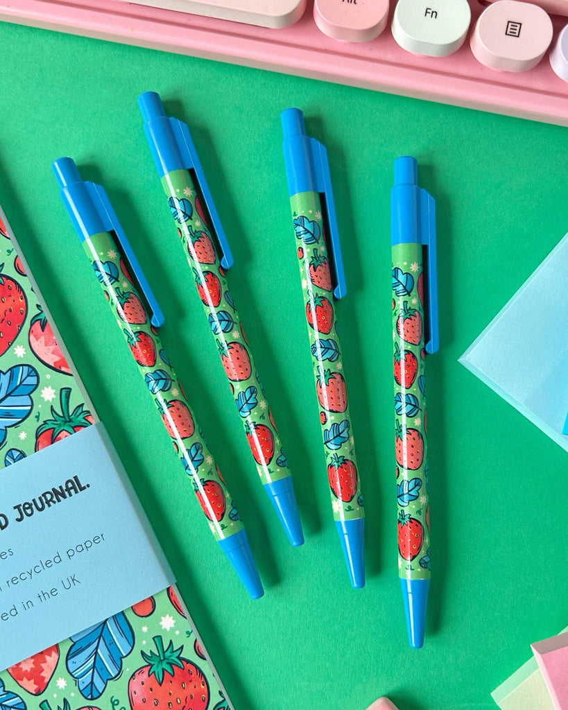 Four Rebel Romance strawberry ballpoint pens laid out on a bright green background alongside a matching strawberry journal and pastel sticky notes. The pens feature a fruity design by Stacey McEvoy Caunt, with blue details and no outer packaging.