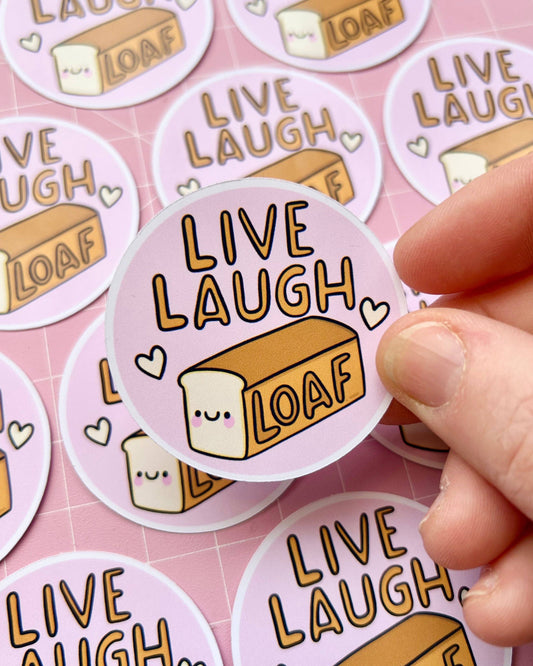A hand holding a cute Rebel Romance glossy sticker designed by Kelly Lou, featuring the words “Live Laugh Loaf” with a smiling loaf of bread and little hearts on a pastel pink background. Perfect for millennials who love alternative style, fun gifts, and quirky illustrations.