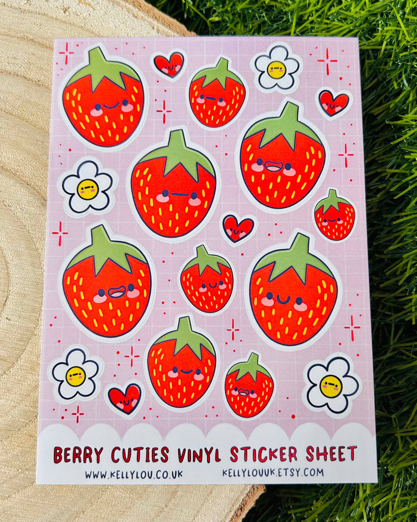 Flat lay of Rebel Romance Berry Cuties Vinyl Sticker Sheet by Kelly Lou, featuring adorable smiling strawberries, heart faces, white daisies, and sparkles on a pink grid background. Printed on waterproof A6 vinyl, perfect for decorating planners, water bottles, or stationery. A sweet and cheerful sticker set ideal for food lovers and fans of kawaii art.