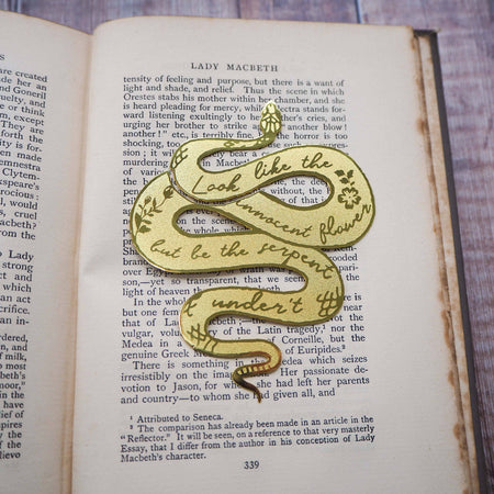 A brass bookmark shaped like a coiled serpent, delicately etched with the famous Lady Macbeth quote: “Look like the innocent flower, But be the serpent under ‘t.” The bookmark is placed inside an open book on a wooden surface, with the title “Lady Macbeth” visible on the page. The snake’s head is designed to peek out from the top of the book when closed, adding a dramatic and literary flair.