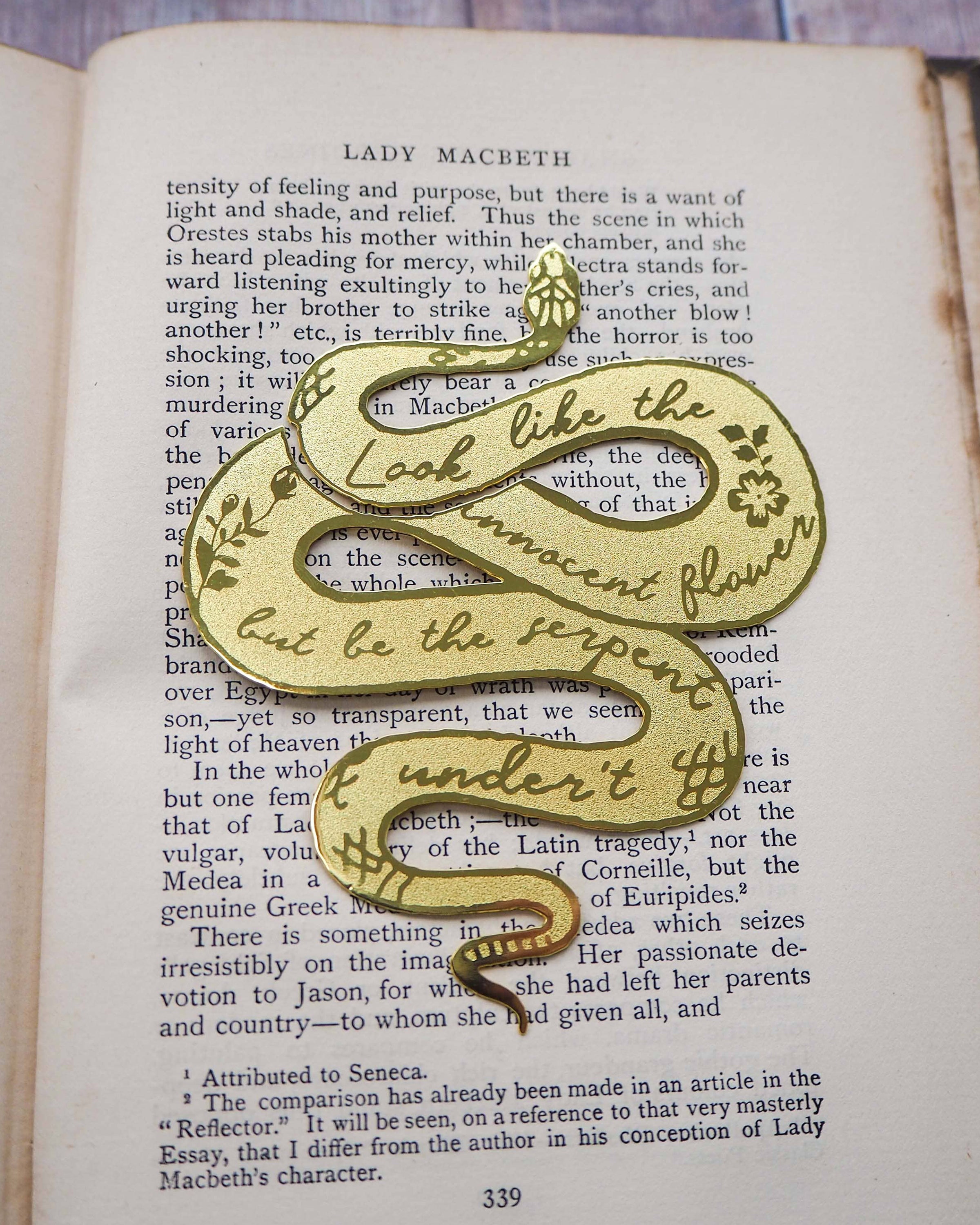 A brass bookmark shaped like a coiled serpent, delicately etched with the famous Lady Macbeth quote: “Look like the innocent flower, But be the serpent under ‘t.” The bookmark is placed inside an open book on a wooden surface, with the title “Lady Macbeth” visible on the page. The snake’s head is designed to peek out from the top of the book when closed, adding a dramatic and literary flair.