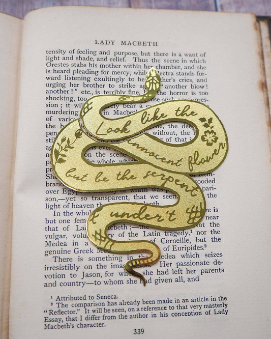 A brass bookmark shaped like a coiled serpent, delicately etched with the famous Lady Macbeth quote: “Look like the innocent flower, But be the serpent under ‘t.” The bookmark is placed inside an open book on a wooden surface, with the title “Lady Macbeth” visible on the page. The snake’s head is designed to peek out from the top of the book when closed, adding a dramatic and literary flair.