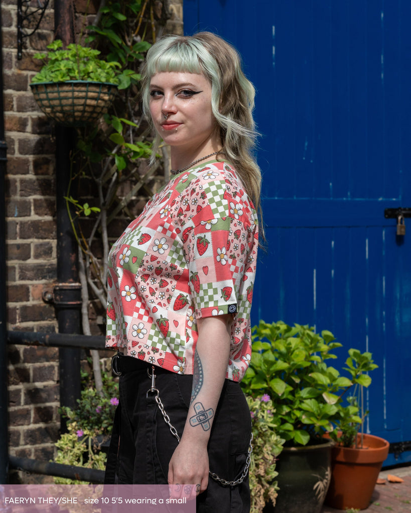 Faeryn, a non binary femme person with dyed green hair and tattoos wearing the strawberry picnic crop top and black cargo trousers. The print features blossoms, strawberries, pink and sage colourblock checkerboard. 