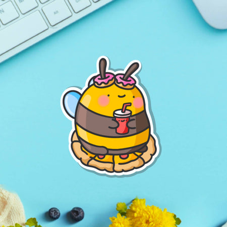 Cute cartoon-style vinyl sticker of a bumblebee with pink doughnut antennae, holding a red drink and sitting on a pie. Ideal for laptops, bottles, and journals.