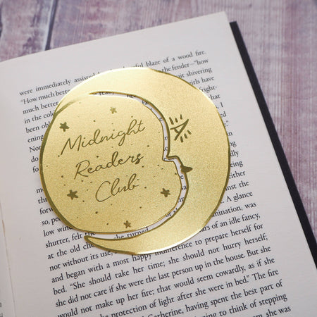 A crescent moon-shaped brass bookmark with “Midnight Readers Club” and celestial stars etched onto its surface, placed on an open book to mark a page. The bookmark adds an elegant and whimsical touch, ideal for book lovers who enjoy night-time reading.