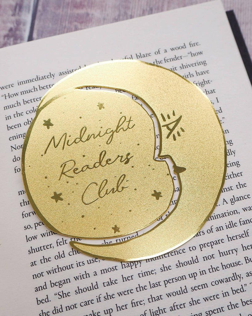 A crescent moon-shaped brass bookmark with “Midnight Readers Club” and celestial stars etched onto its surface, placed on an open book to mark a page. The bookmark adds an elegant and whimsical touch, ideal for book lovers who enjoy night-time reading.