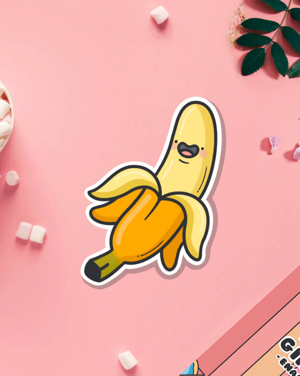 Cute banana vinyl sticker by Toastedink, sold by Rebel Romance, featuring a cartoon banana with a happy face on a pastel pink background, surrounded by mini marshmallows and petals.