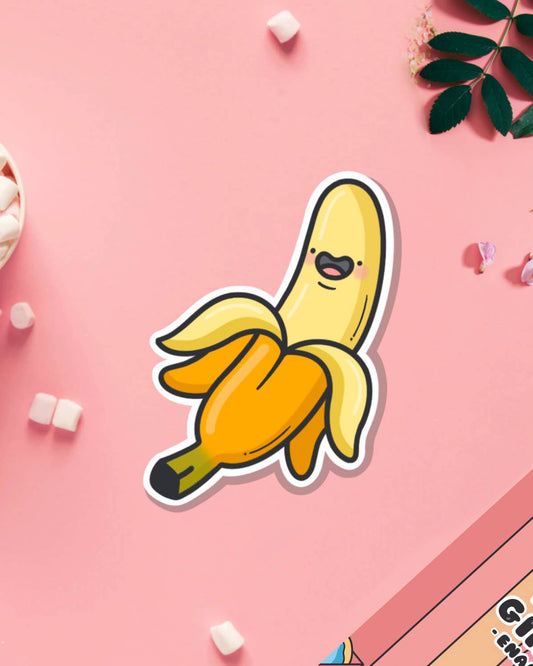 Cute banana vinyl sticker by Toastedink, sold by Rebel Romance, featuring a cartoon banana with a happy face on a pastel pink background, surrounded by mini marshmallows and petals.