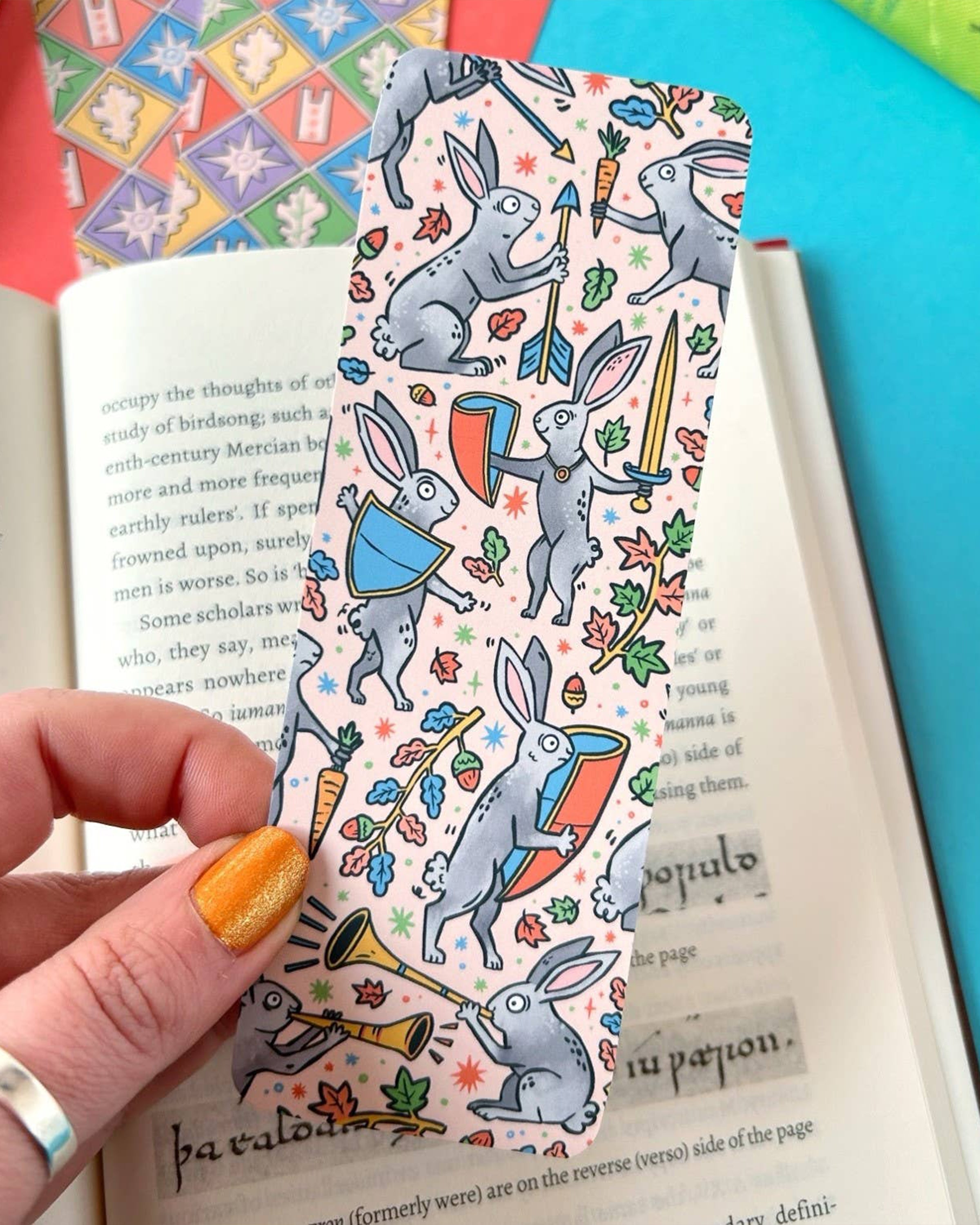 Hand holding the Rebel Romance Medieval Rabbits Bookmark with a view of both its front illustration of battle-ready bunnies and the back’s geometric tile design. Displayed on top of an open book with other bookmarks and books in the background.