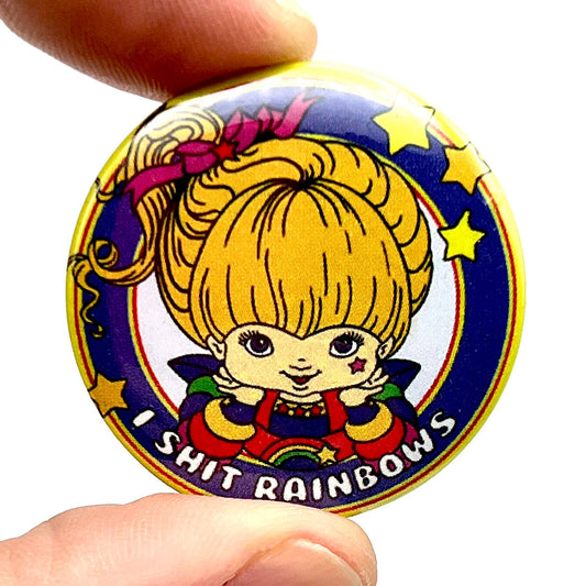 Close-up of a colourful Rebel Romance button pin badge featuring a retro-style cartoon girl with blonde hair, stars, and a rainbow outfit. The text reads “I Shit Rainbows” in bold white letters on a purple background. Designed by Bite Your Granny and held between two fingers. Quirky, cheeky gift for fans of alternative fashion and bold humour.