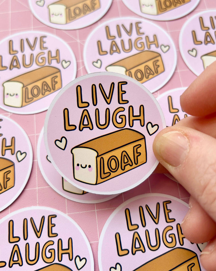 A hand picking up a Rebel Romance “Live Laugh Loaf” glossy sticker by Kelly Lou, surrounded by more stickers on a pink background. The design features a cute illustrated loaf and hearts, ideal for alternative fashion fans and gift lovers.