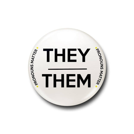 A 25mm white lapel pin featuring bold black text that reads “They/Them,” with “Pronouns Matter” written in smaller curved text around the edge, accompanied by small star accents.