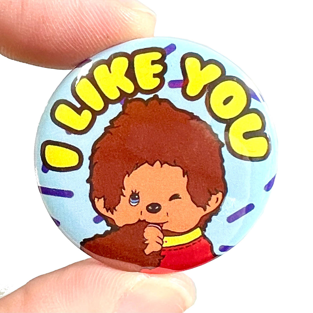 A close-up of a round button pin badge being held between two fingers. The badge features a retro-style illustration of a brown fuzzy character inspired by Chick-A-Boo, winking and sucking its thumb, with the bold yellow text “I LIKE YOU” above. Designed by Bite Your Granny, sold by Rebel Romance, a UK brand for alternative fashion and nostalgic gifts.