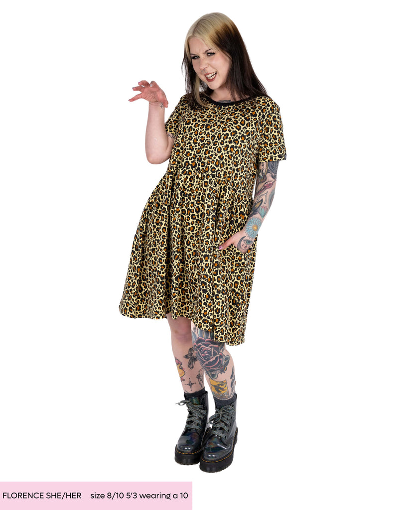 Florence, alternative tattooed woman with dyed blonde and black hair, posing with her hand up and pulling a growling type of face wearing the rebel romance leopard print smock dress. She is wearing the natural leopard coloured dress with grey sparkly boots in a white studio background.