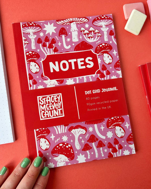 Person with green painted nails holds the Stacey McEvoy Caunt pink and red mushroom dot grid journal sold by Rebel Romance. The notebook cover features magical red and white mushroom illustrations on a pink background.