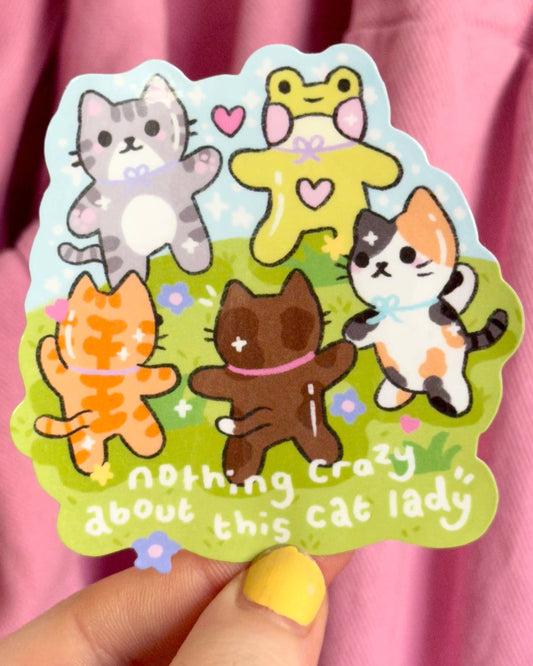 Glossy vinyl cat sticker held in front of a pink fabric background, featuring cute illustrated cats and a frog dancing with the phrase “nothing crazy about this cat lady.” Fun, pastel sketch-style design from Rebel Romance, a small UK brand that celebrates alternative fashion and gifts for millennials.