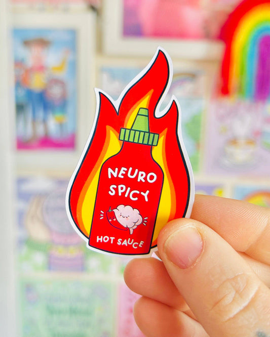 A hand holds a glossy vinyl sticker shaped like a bottle of hot sauce surrounded by cartoon flames. The red bottle features the text "Neuro Spicy Hot Sauce" and a cute smiling brain hugging a chilli pepper. Designed by Kellylou, this vibrant sticker is photographed in front of a colourful, rainbow-themed background. Sold by Rebel Romance, a UK small business that celebrates alternative fashion and neurodivergent pride.
