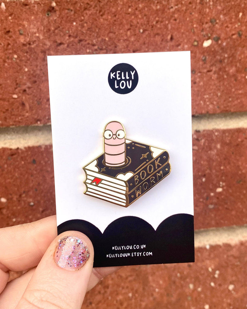 A hand holds up the Kelly Lou Bookworm Enamel Pin against a red brick wall. The gold pin shows a pink worm with glasses sitting on a black and gold book marked "Book Worm", attached to a Rebel Romance backing card. Sold by Rebel Romance, a UK indie brand specialising in alternative style, quirky gifts, and slow ethical fashion.
