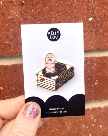 A hand holds up the Kelly Lou Bookworm Enamel Pin against a red brick wall. The gold pin shows a pink worm with glasses sitting on a black and gold book marked "Book Worm", attached to a Rebel Romance backing card. Sold by Rebel Romance, a UK indie brand specialising in alternative style, quirky gifts, and slow ethical fashion.