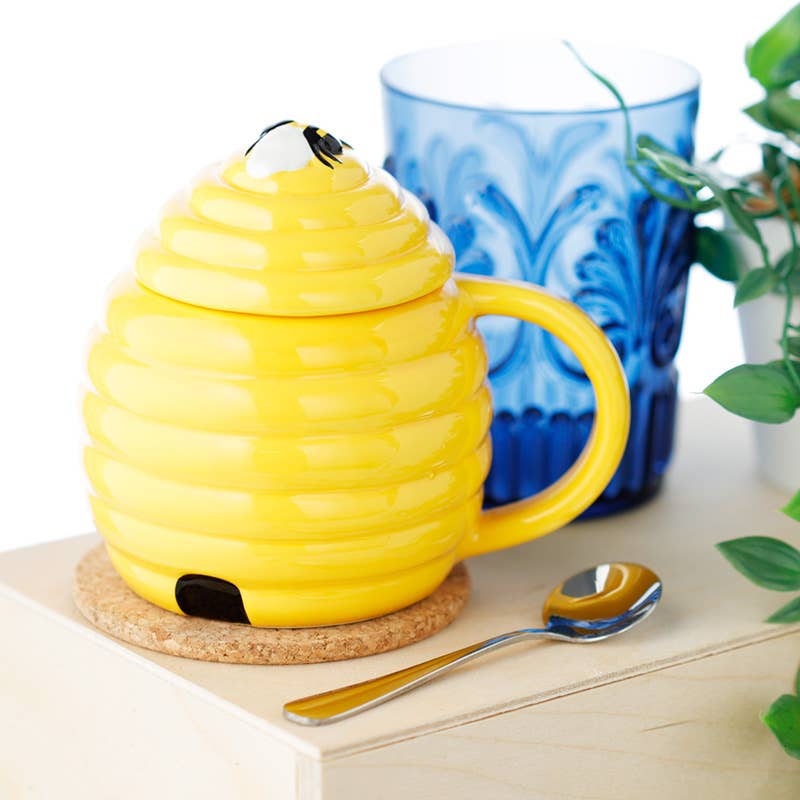 The beehive mug with its lid on, displayed on a cork coaster next to a blue glass and spoon. A whimsical ceramic mug from Rebel Romance, ideal for tea lovers and gift-givers.