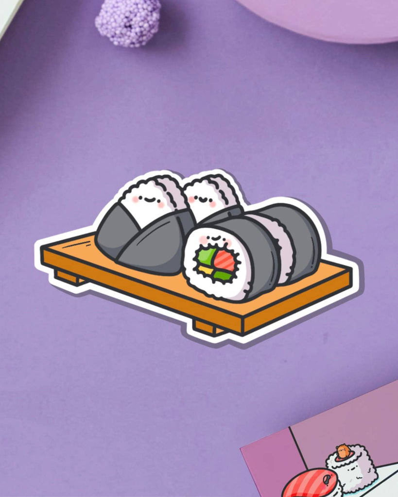Adorable sushi-themed vinyl sticker showing cartoon sushi rolls and onigiri with smiling faces on a wooden tray. Displayed on a purple backdrop with textured props. Available at Rebel Romance, a UK-based store for quirky, cute and alternative millennial aesthetics.