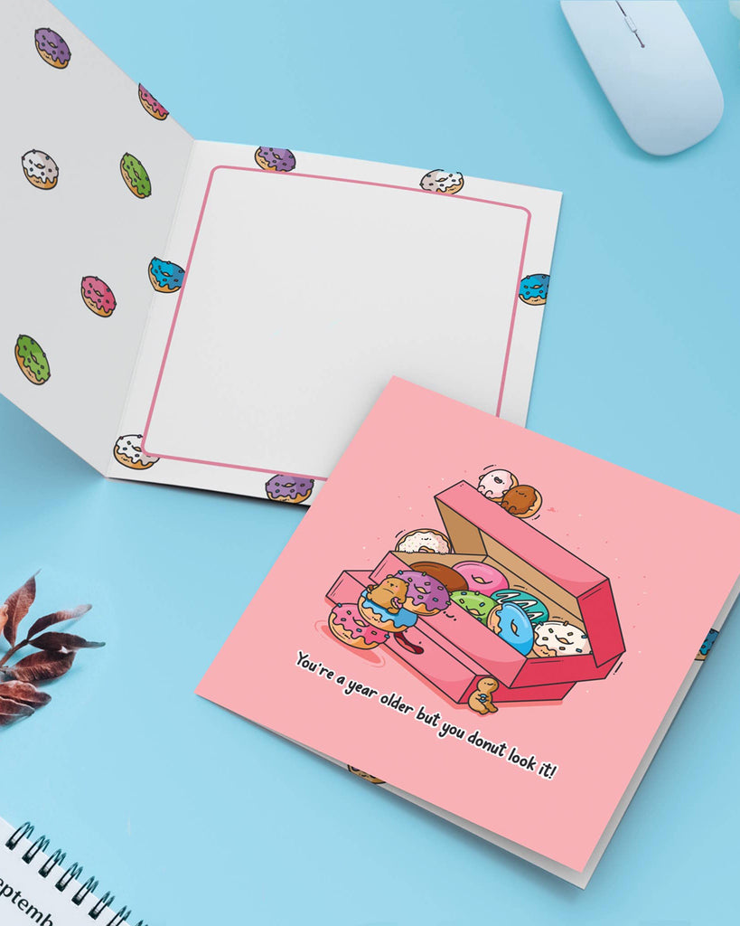 Open Toastedink donut birthday card on a flat lay with a blue background, revealing the donut-printed interior and the pun-filled pink card cover saying "You're a year older but you donut look it!"