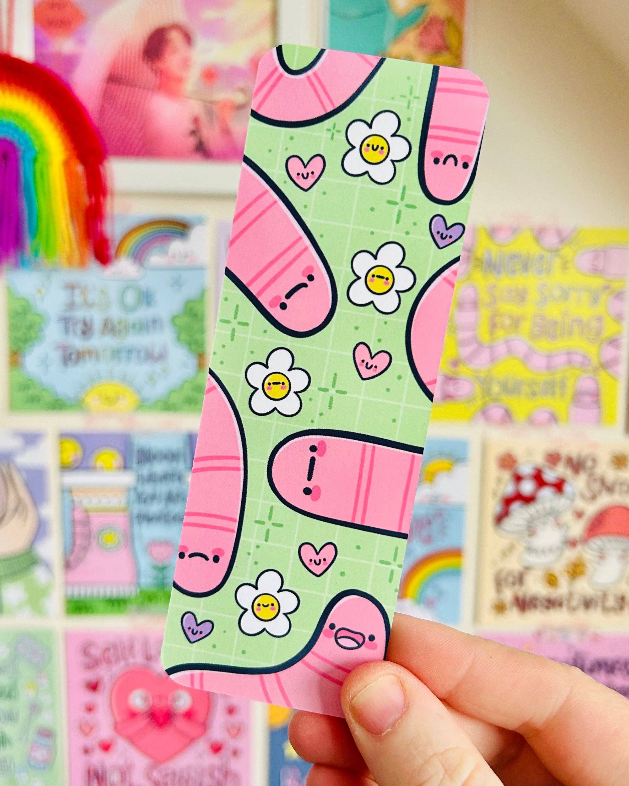 Hand holding the Wormies Bookmark by Kelly Lou from Rebel Romance in front of a colourful wall of art prints. The bookmark features pink worms, happy daisies and tiny hearts on a green grid design. A vibrant, illustrated accessory from a UK brand celebrating alternative style, playful art and millennial‑friendly gifts.