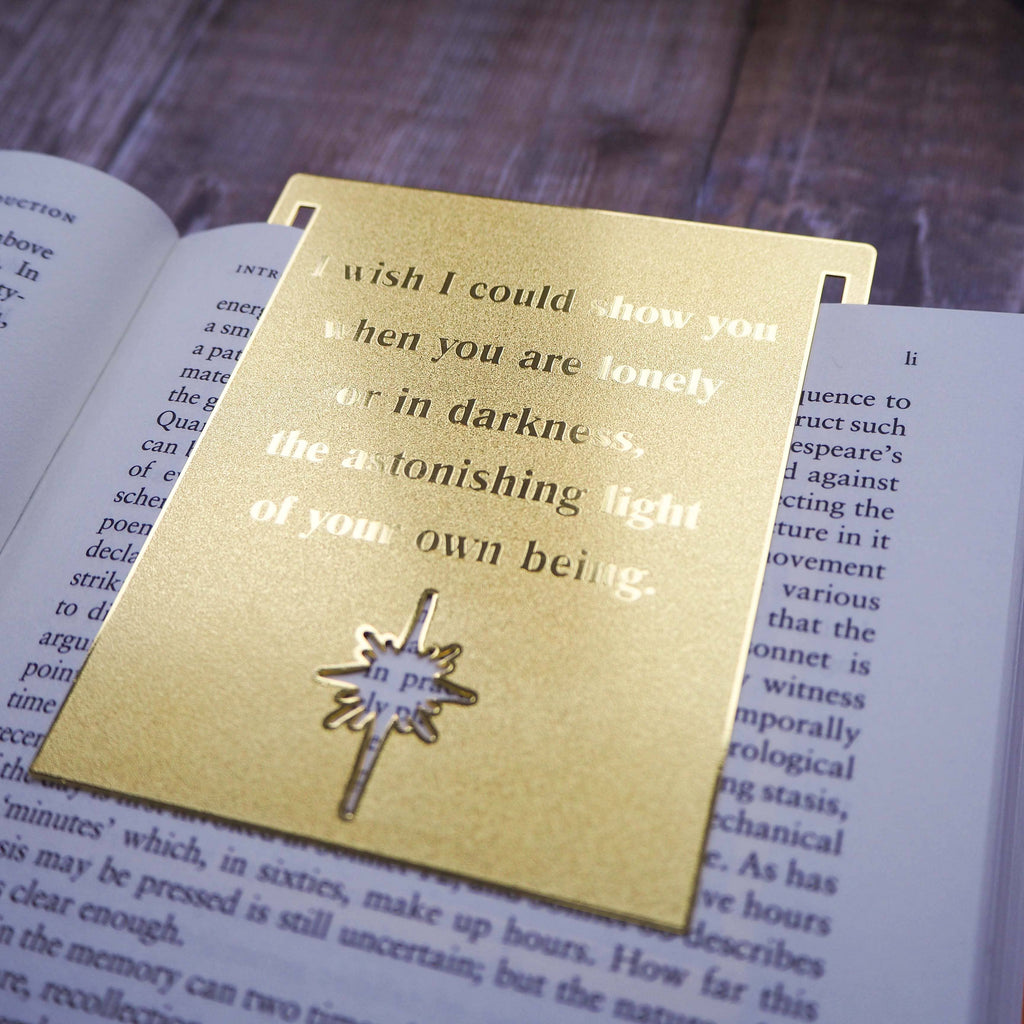 A close-up image of a brass bookmark engraved with the heartfelt Hafiz of Shiraz quote: “I wish I could show you, when you are lonely or in darkness, the astonishing light of your own being.” The bookmark is resting on the pages of an open book, showcasing its intricate detailing and elegant design.