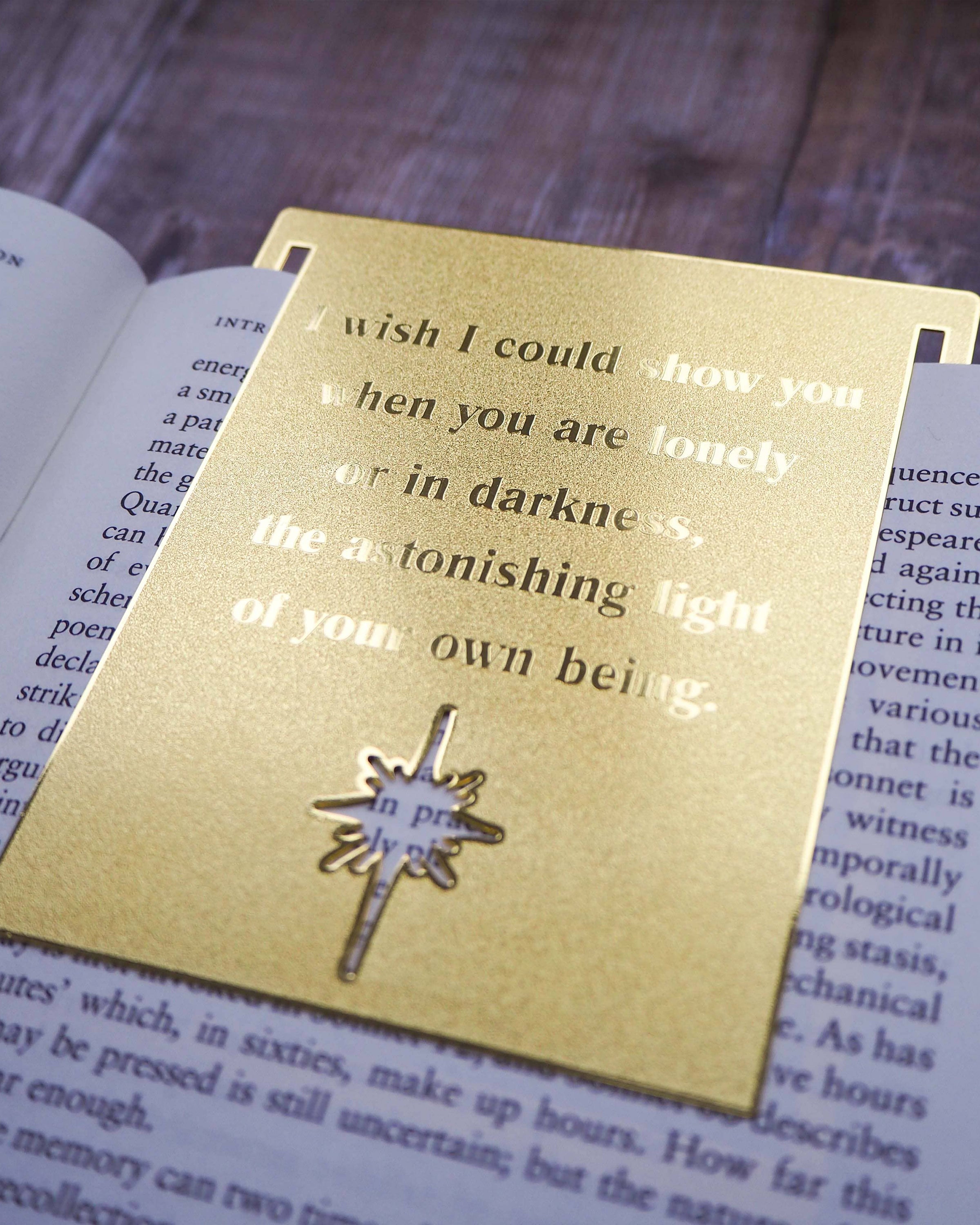 A close-up image of a brass bookmark engraved with the heartfelt Hafiz of Shiraz quote: “I wish I could show you, when you are lonely or in darkness, the astonishing light of your own being.” The bookmark is resting on the pages of an open book, showcasing its intricate detailing and elegant design.