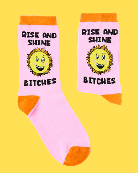 Flat lay of novelty sunshine socks on a bright yellow background, featuring cheerful sun faces and the phrase "Rise and Shine Bitches." Sold by Rebel Romance, a UK small biz offering bold, alternative fashion and retro vibes.