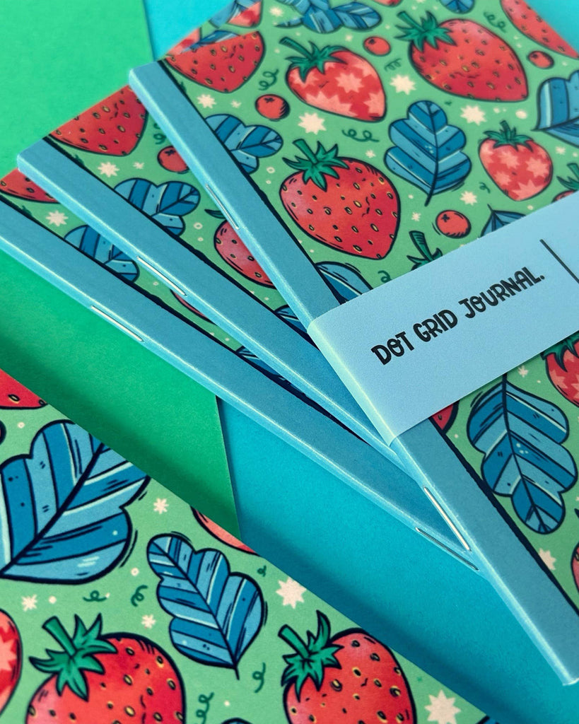 Stack of Rebel Romance Strawberry Pocket Notebooks arranged diagonally, displaying their colourful strawberry covers and neat square spines. A branded bellyband highlights its recycled paper, UK printing, and dot grid interior – perfect for stylish stationery lovers.