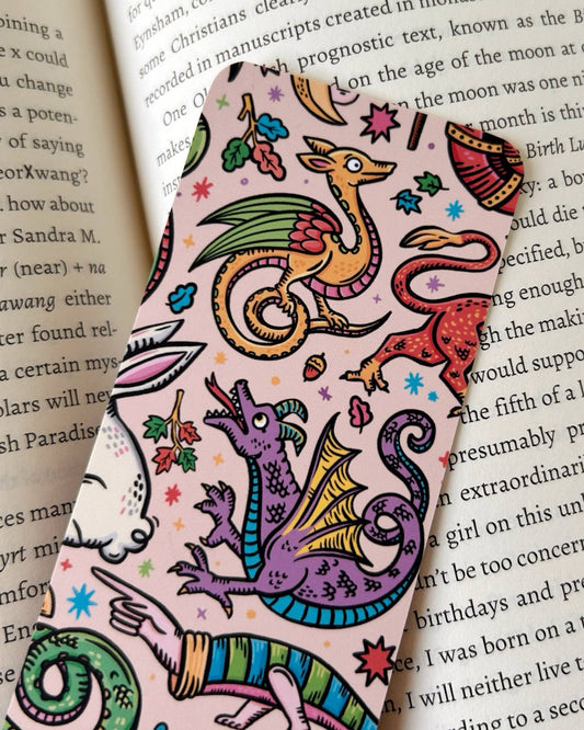 A close-up of a colourful medieval creatures bookmark by Stacey McEvoy Caunt, featuring dragons, moons, and leaves on a pastel background, peeking out from the pages of a book. Sold by Rebel Romance, a small UK brand celebrating alternative and whimsical style.