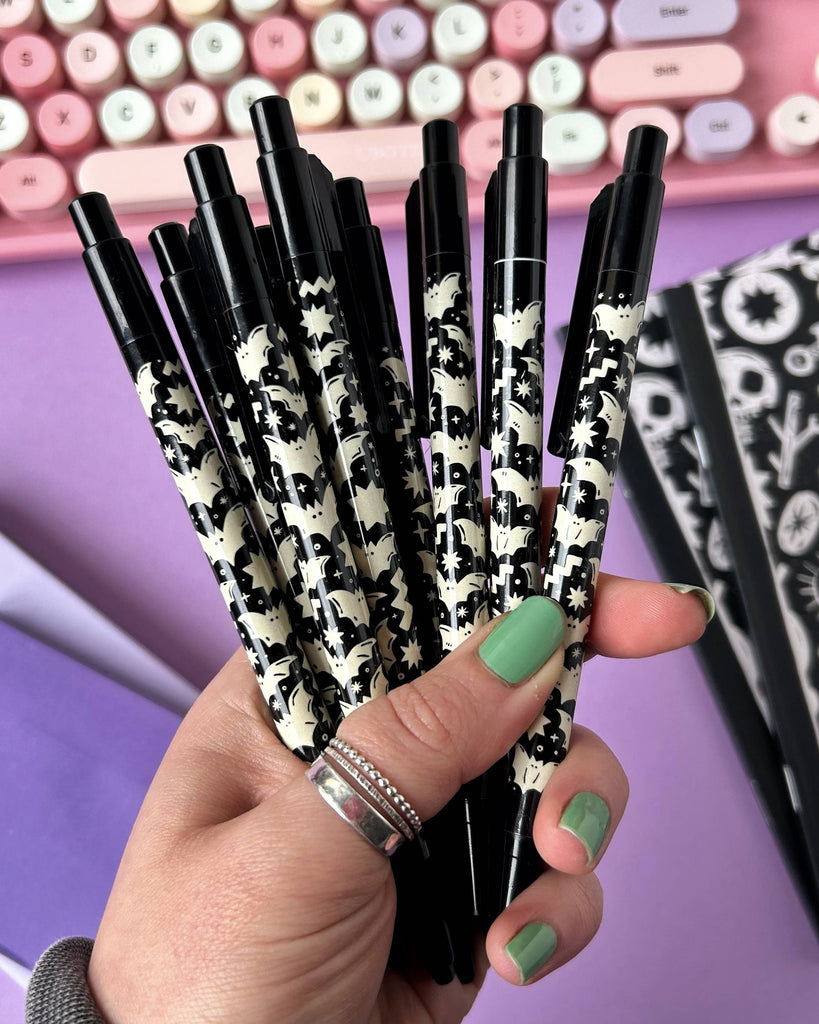 Hand holding a fan of eight Rebel Romance Bat Pattern Pens against a purple background, showing the repeated black and white bat and star pattern and a pastel keyboard.