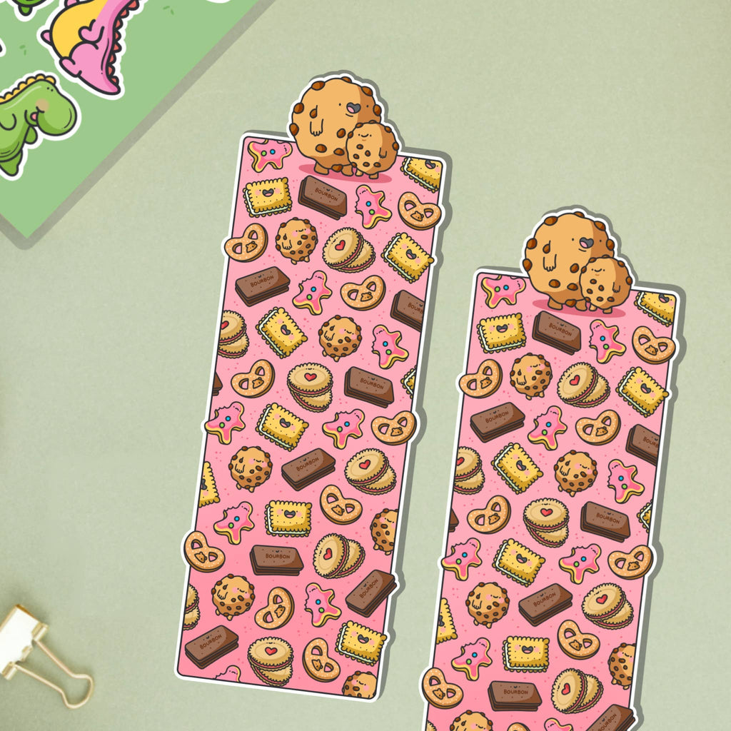 Glossy biscuit-themed bookmark featuring cartoon illustrations of cookies, pink iced biscuits, bourbons, jammy dodgers and other treats on a pink background. Topped with two cute chocolate chip cookies.
